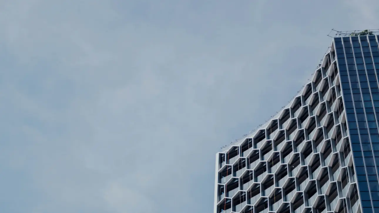 Uma fotografia em ângulo baixo de um arranha-céu moderno com uma fachada geométrica de colmeia hexagonal intrincada e uma seção de vidro estreita, focado no canto direito. O céu nublado preenche o restante da imagem. A imagem serve para ilustrar 'Como o processo de compra de imóvel mudou com a chegada do digital'.