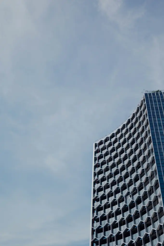 Uma fotografia em ângulo baixo de um arranha-céu moderno com uma fachada geométrica de colmeia hexagonal intrincada e uma seção de vidro estreita, focado no canto direito. O céu nublado preenche o restante da imagem. A imagem serve para ilustrar 'Como o processo de compra de imóvel mudou com a chegada do digital'.