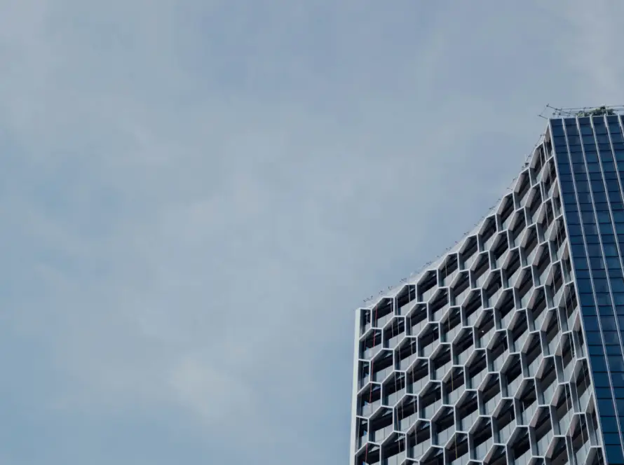 Uma fotografia em ângulo baixo de um arranha-céu moderno com uma fachada geométrica de colmeia hexagonal intrincada e uma seção de vidro estreita, focado no canto direito. O céu nublado preenche o restante da imagem. A imagem serve para ilustrar 'Como o processo de compra de imóvel mudou com a chegada do digital'.