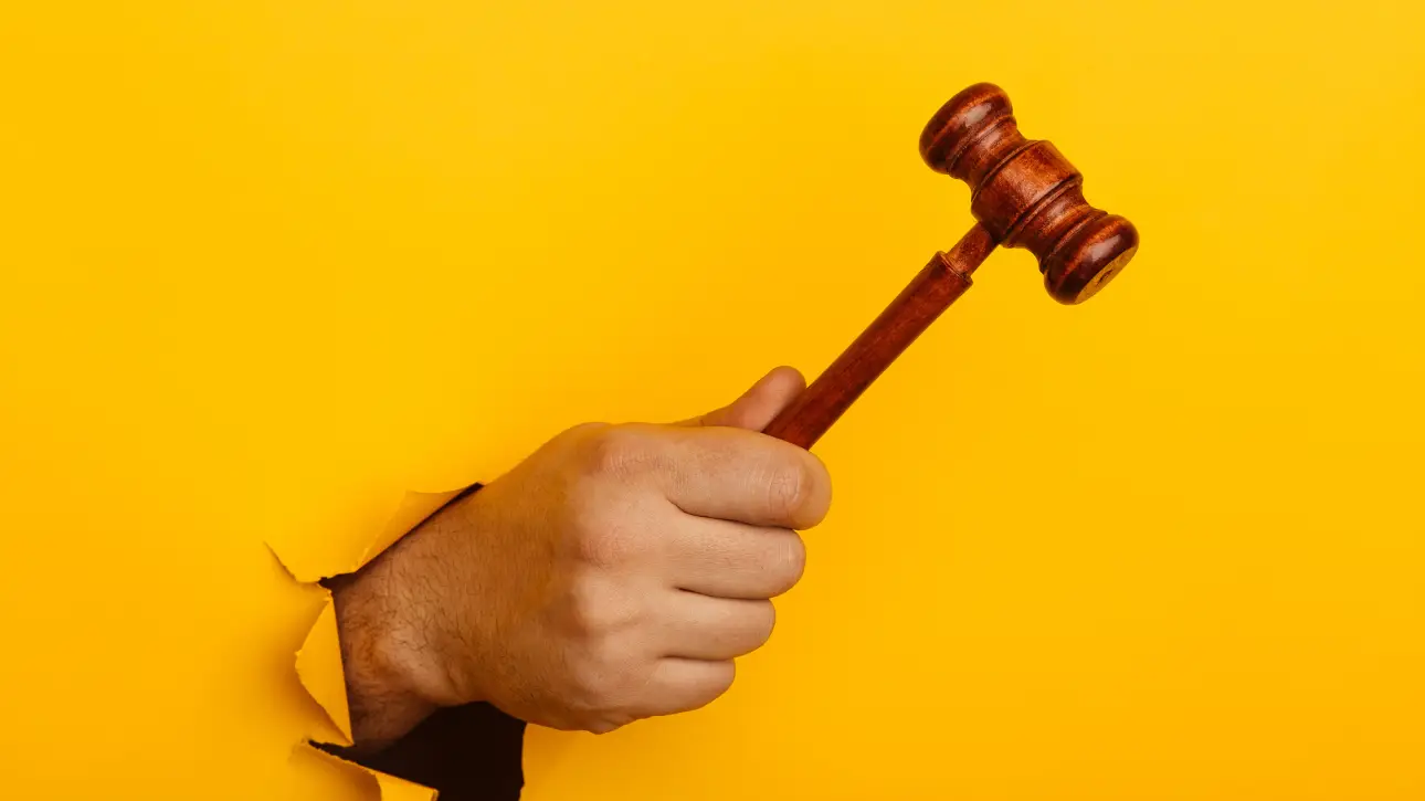 mão segurando um martelo de juiz através de um rasgo em fundo amarelo, representando o leilão de anúncios e como funciona no Meta Ads